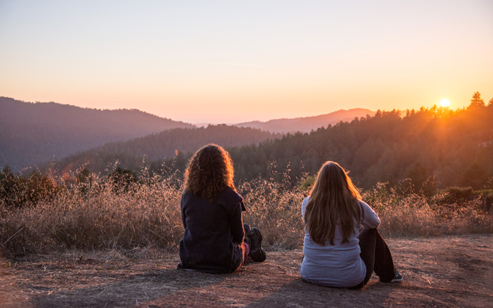 Two friends sit on the trail, observing the sun setting behind a tree-covered ridgeline.
