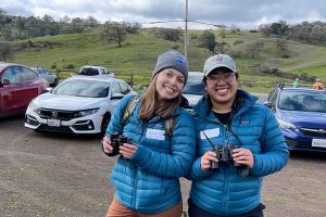 A pair of POST staff members at a birding event wear matching puffy jackets and binoculars.