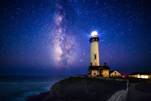Pigeon Point Lighthouse sits on a cliff's edge overlooking the ocean, framed by the starry night sky and Milky Way.